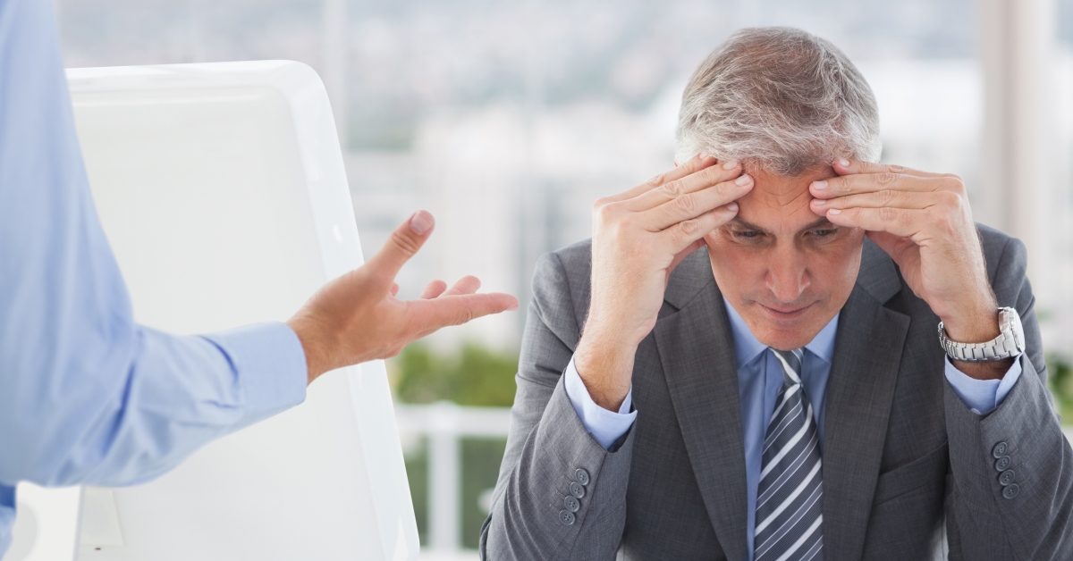 Frustrated businessman sitting with hands on forehead computer literature environment thoughtful sophisticated 1200X628